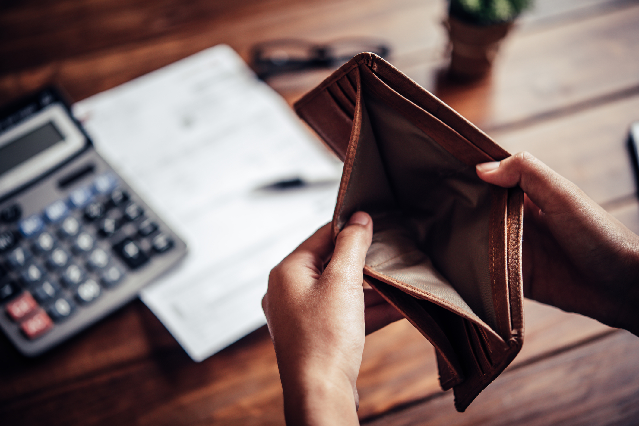 An open, empty wallet held by two hands is pictured in front of a wooden desk with a calculator, papers, and glasses