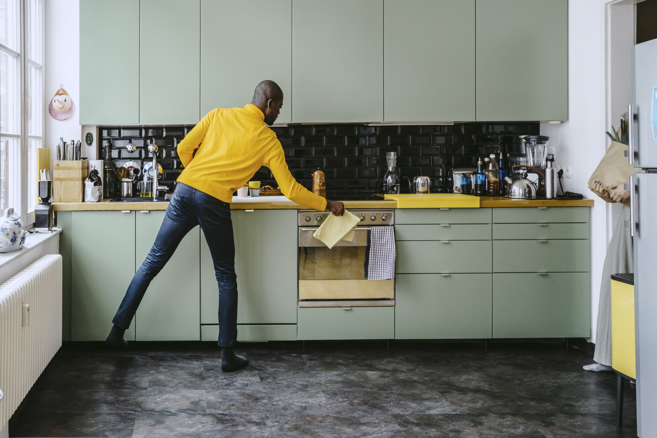 A person in a yellow jumper and dark jeans tidying a modern kitchen with light green cabinets and black backsplash. They hold a yellow cloth near the oven