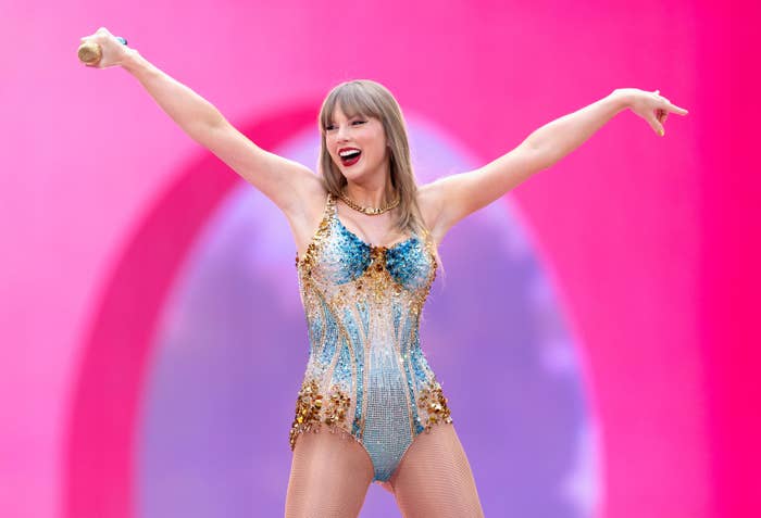Taylor Swift performs on stage, wearing a sparkling bejeweled bodysuit, holding a microphone and smiling with arms raised