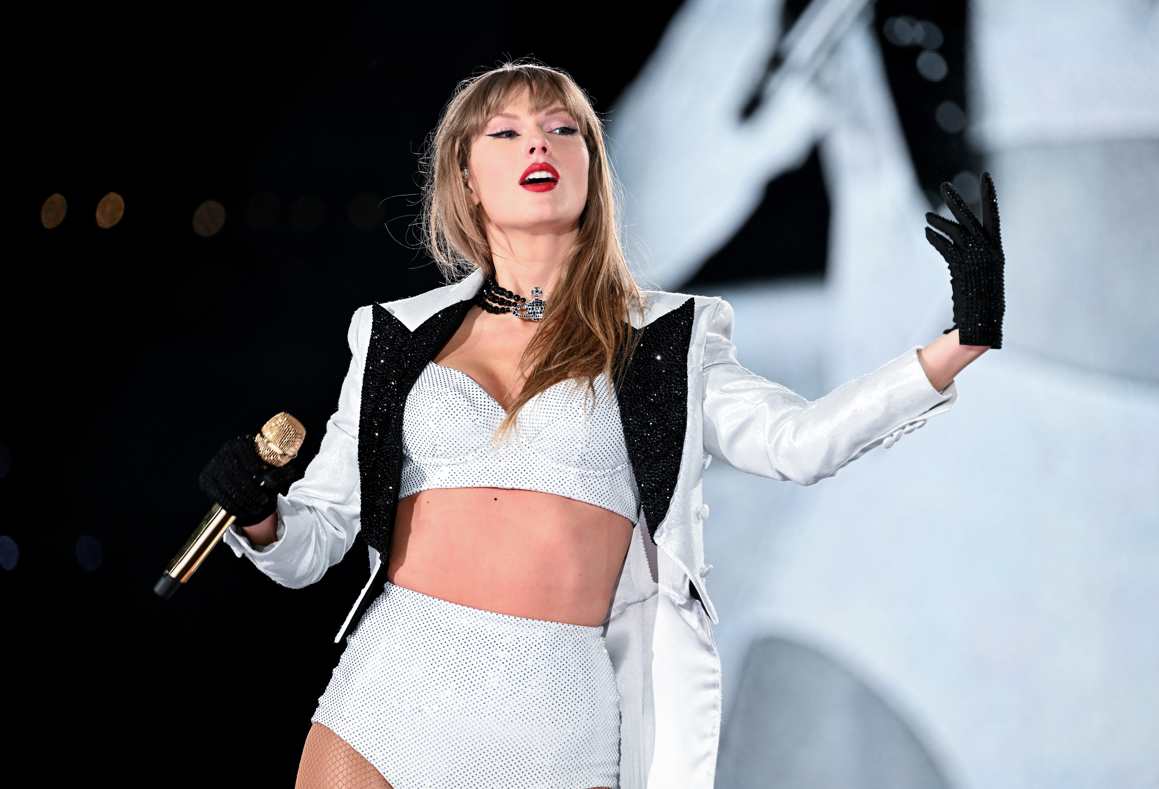 Taylor Swift performs on stage wearing a crop top and high-waisted shorts with a blazer and black gloves. She holds a gold microphone