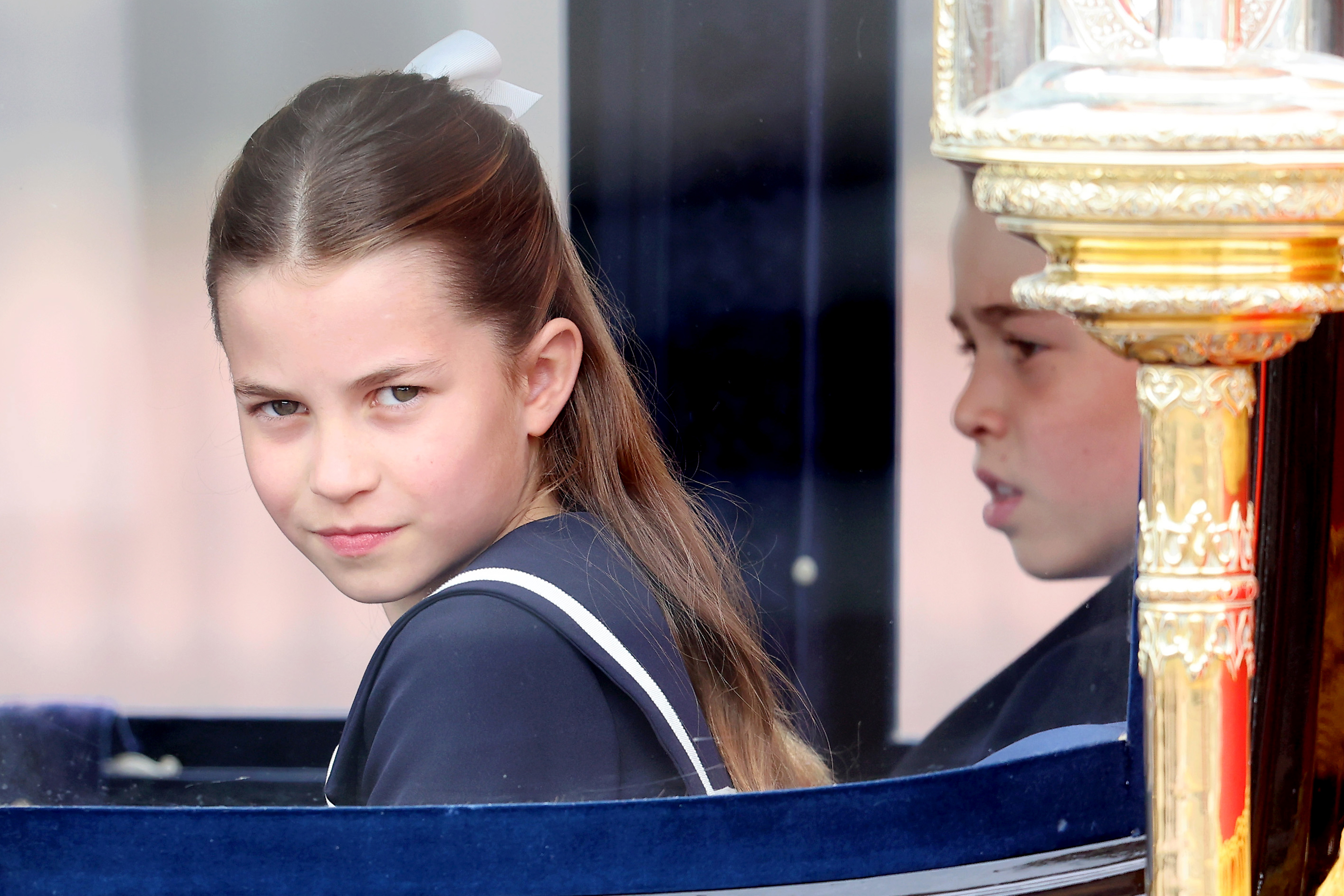 Princess Charlotte dressed in a formal outfit with a white bow in her hair, sitting in a carriage, with her reflection visible in the window