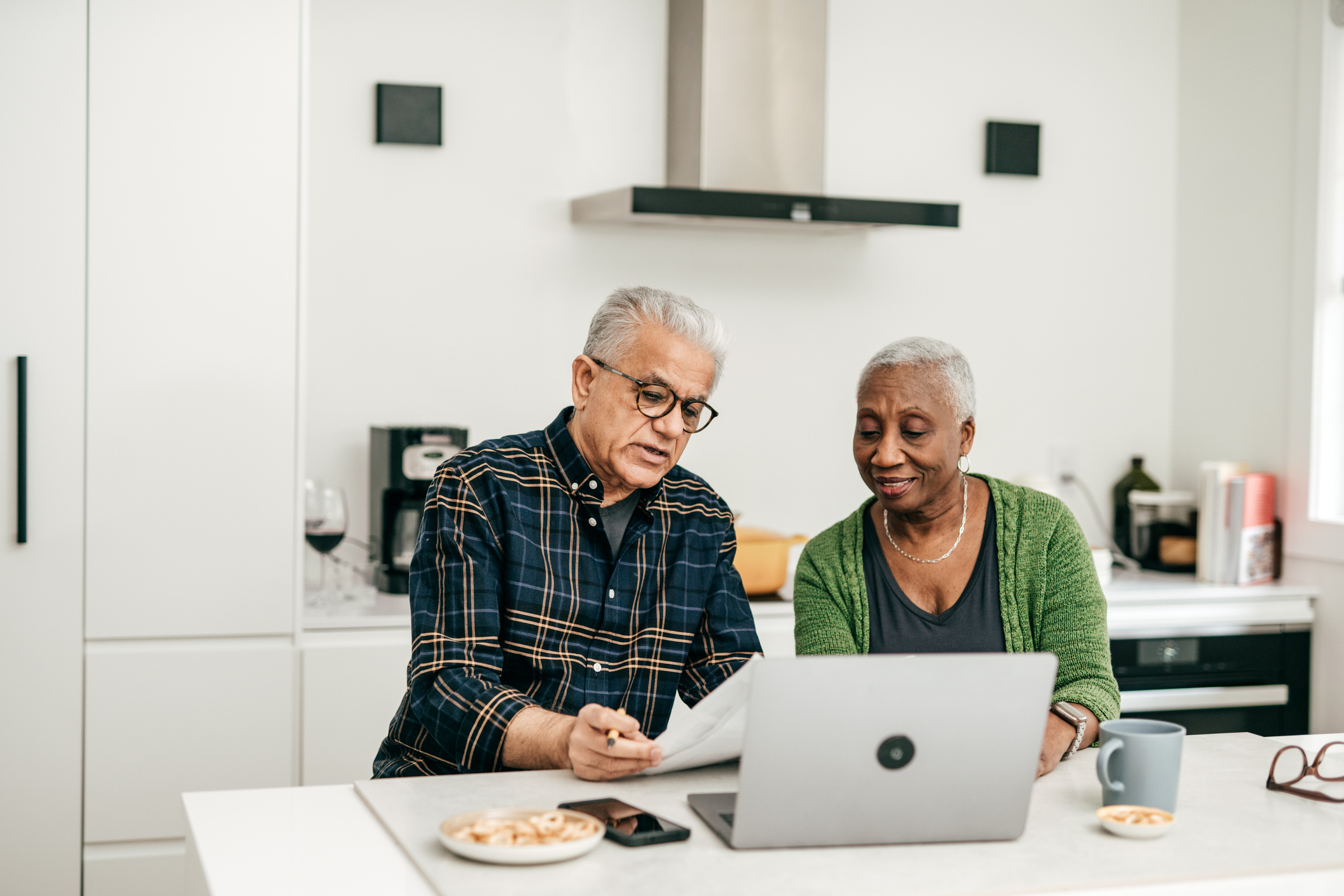 An elderly man and woman sit at a kitchen counter, reviewing documents and using a laptop, possibly discussing finances or work-related matters