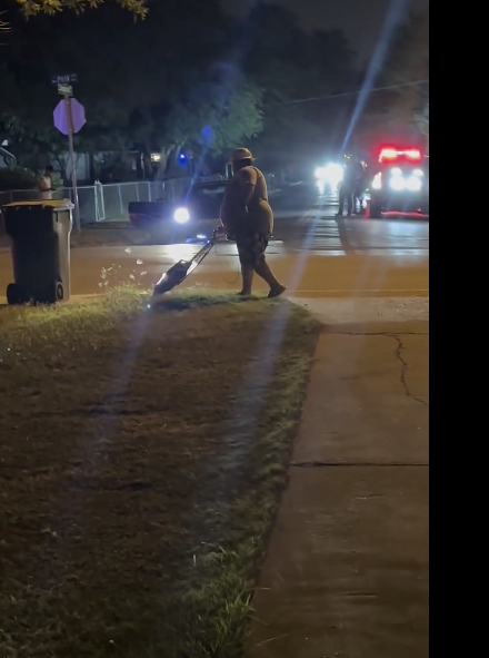 A person is sweeping leaves off the sidewalk at night in a suburban area. Cars with headlights on and one with flashing lights are visible in the background