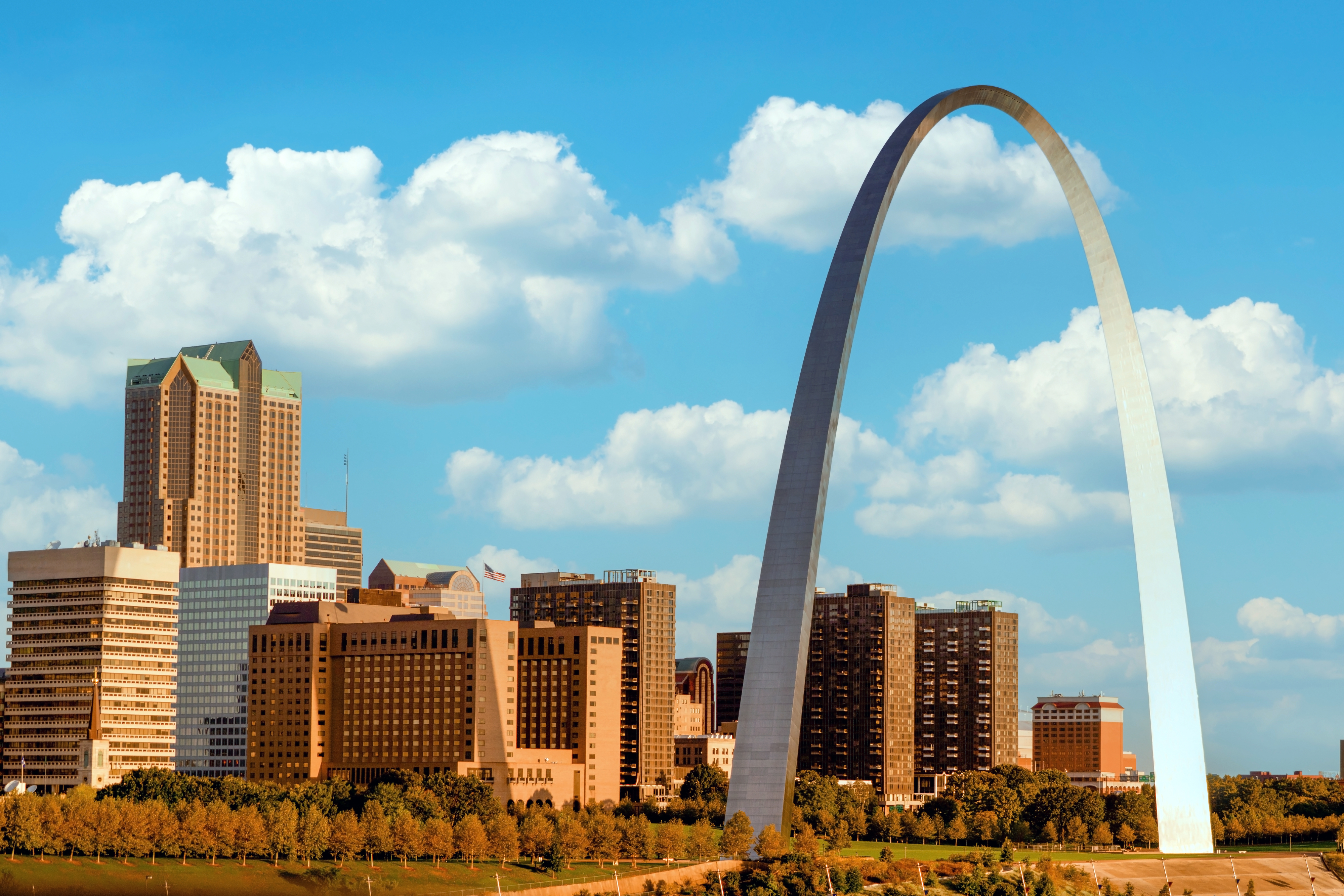 The image shows the Gateway Arch in St. Louis, with a cityscape of tall buildings and a partly cloudy sky in the background