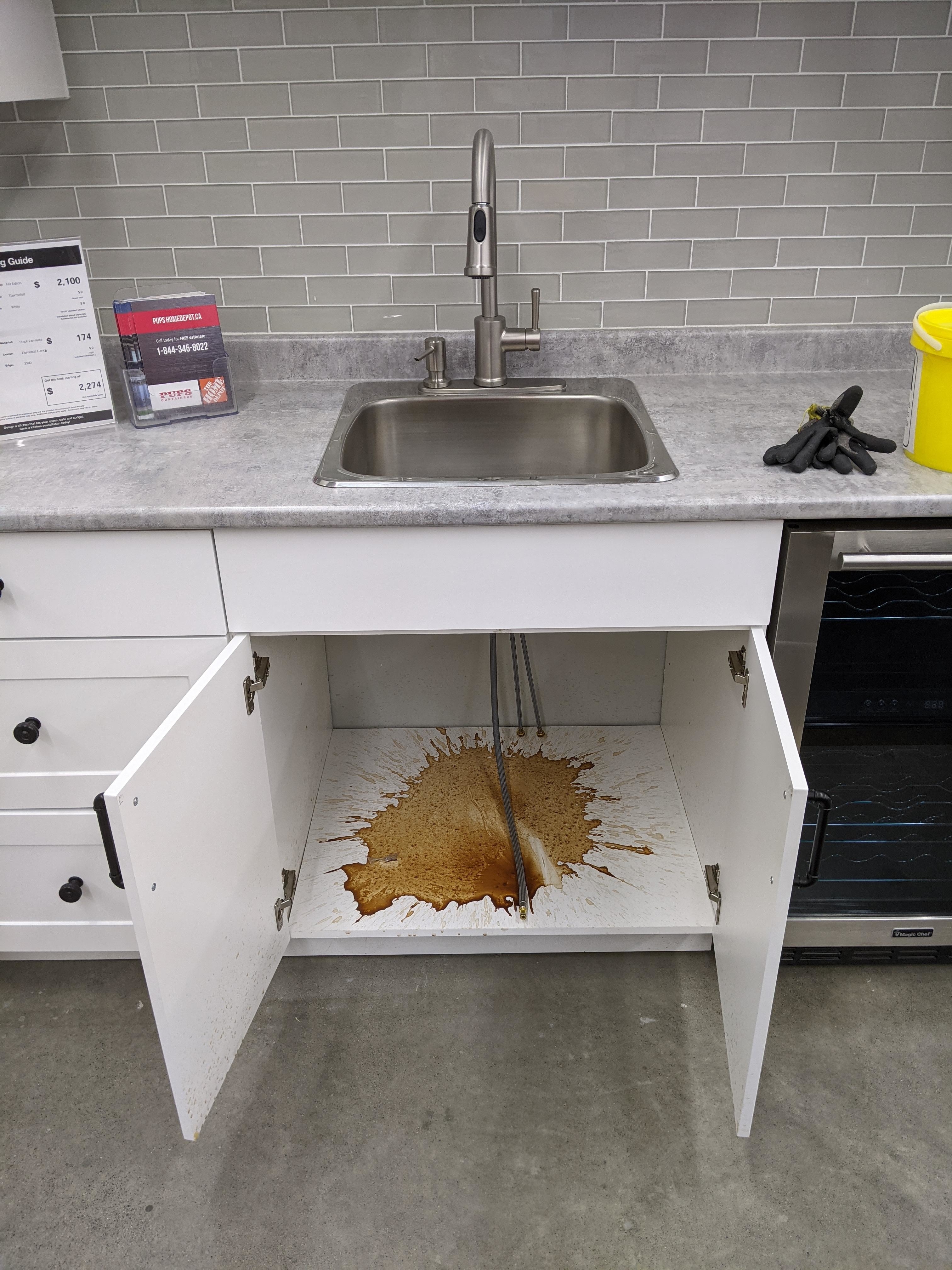 Sink overflows with brown liquid into an open cabinet below, creating a large spill. Countertop items include a box, towels, and a yellow bucket on the right