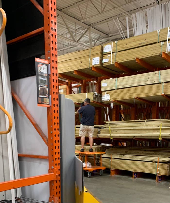 A person stands on a platform lifting wood boards in a hardware store aisle filled with stacked lumber