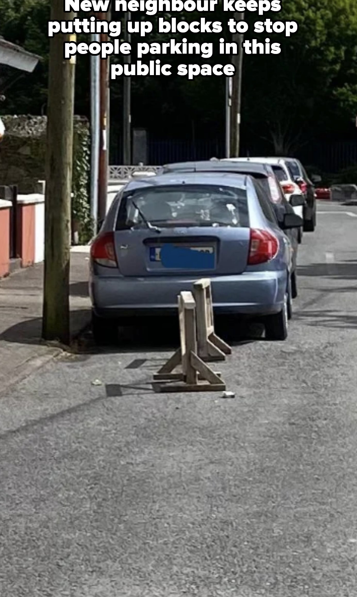 A silver car is parked in front of a wooden barrier on a residential street. Power lines and trees are visible in the background