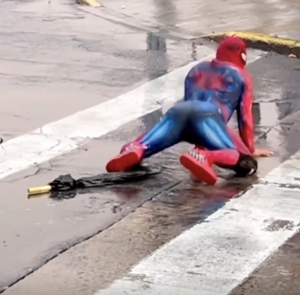 A person in a Spider-Man costume is sprawled on a wet crosswalk with an umbrella nearby