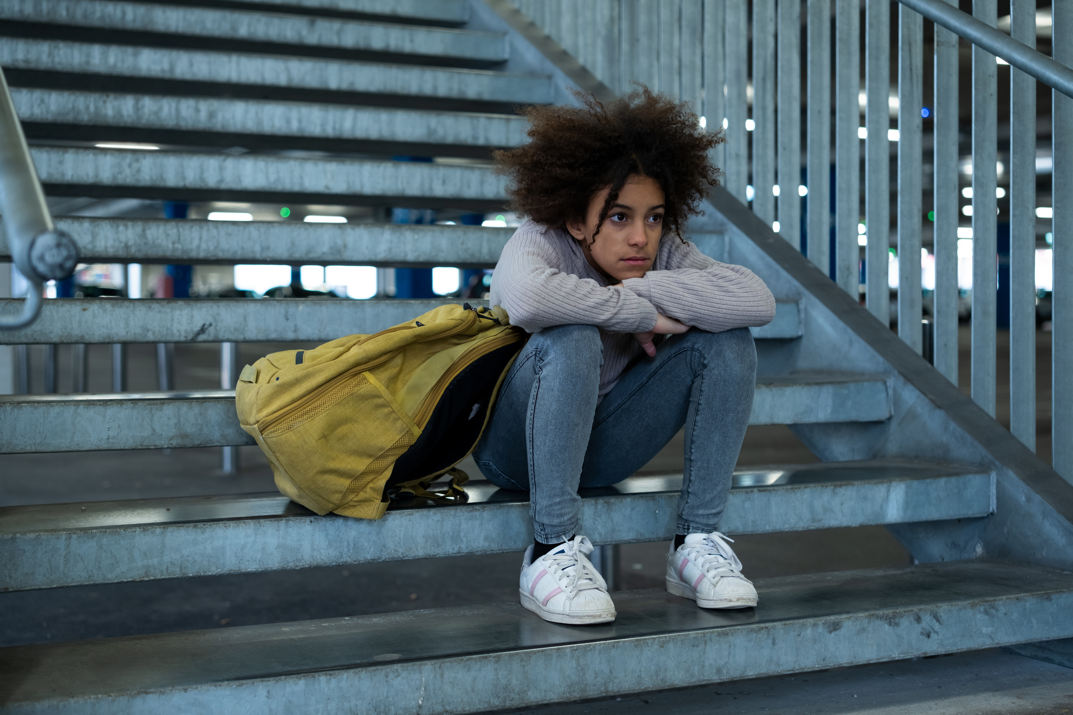 Person sitting on a stairway with a yellow backpack beside them, looking thoughtful and distant