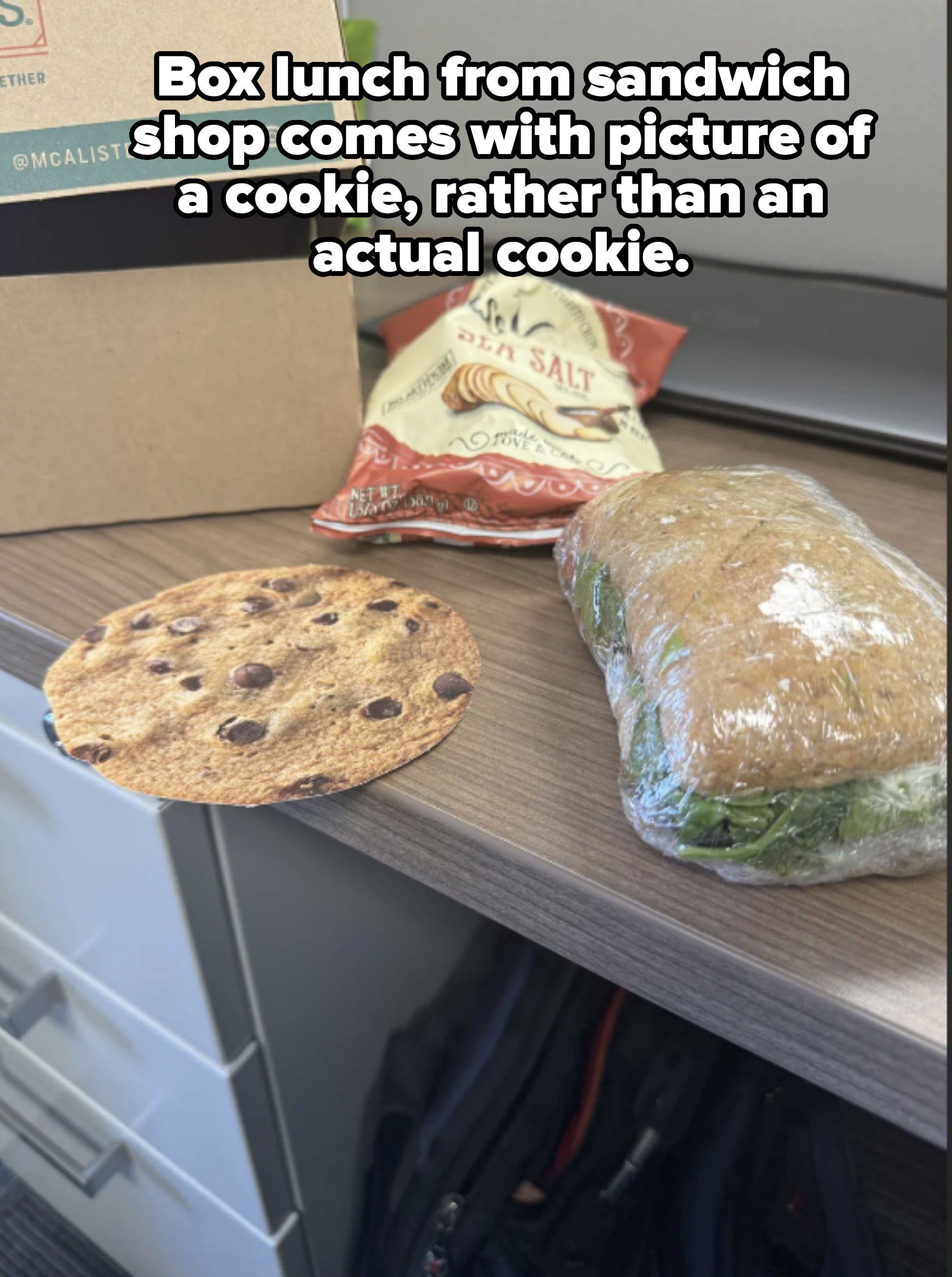 A cookie with chocolate chips, a bag of sea salt chips, and a wrapped sandwich on a desk with a laptop in the background