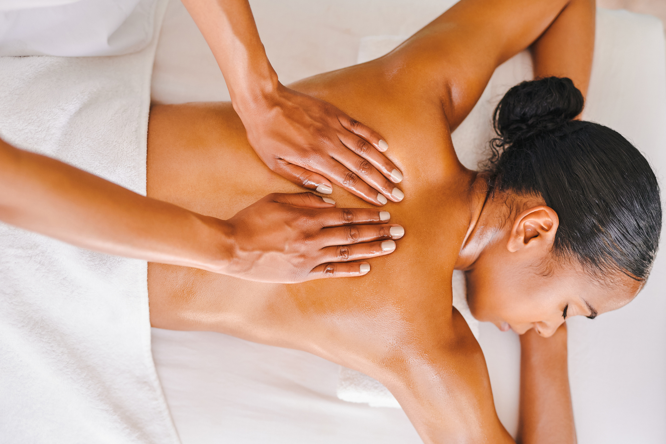 Person receiving a back massage while lying face down on a table covered with a white towel. Hands are shown massaging the person&#x27;s back