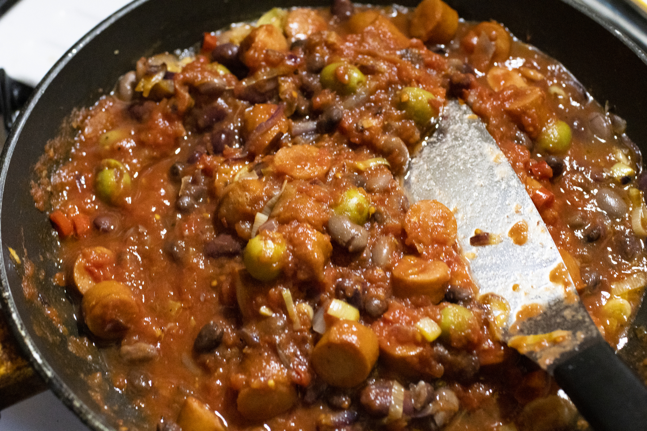 A pan filled with a chunky tomato-based stew featuring olives, sausage slices, and various vegetables, with a spatula resting on the side