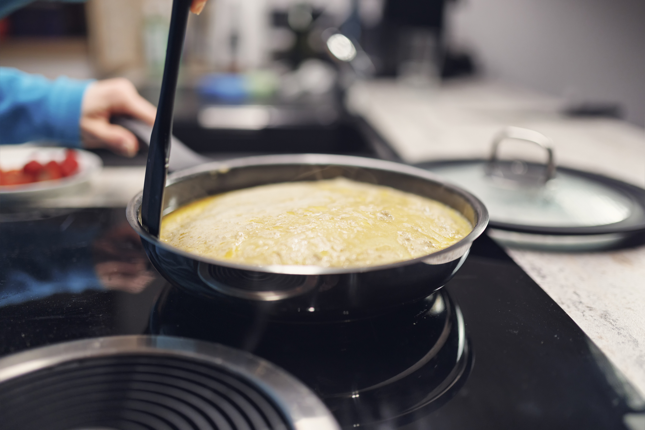 A hand stirs a simmering yellow sauce in a pan on a stovetop. In the background, there are a bowl of cherry tomatoes and a pot lid