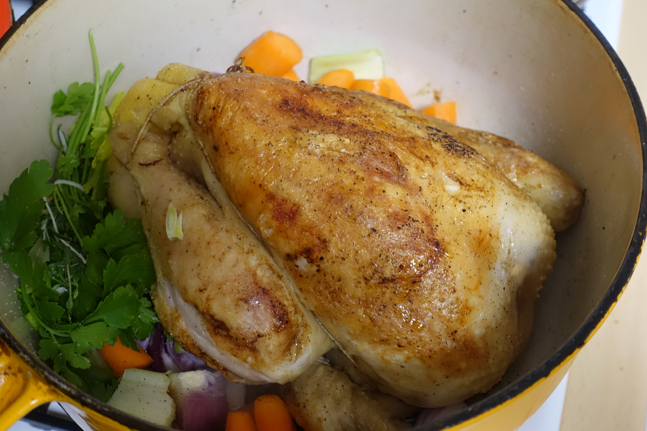 A roasted chicken sits in a pot with chopped vegetables such as carrots, onions, and herbs