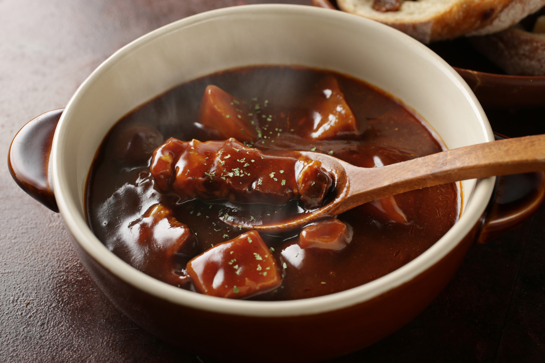 A close-up of a bowl of beef stew with chunks of meat and vegetables, garnished with herbs, and a wooden spoon lifting a piece