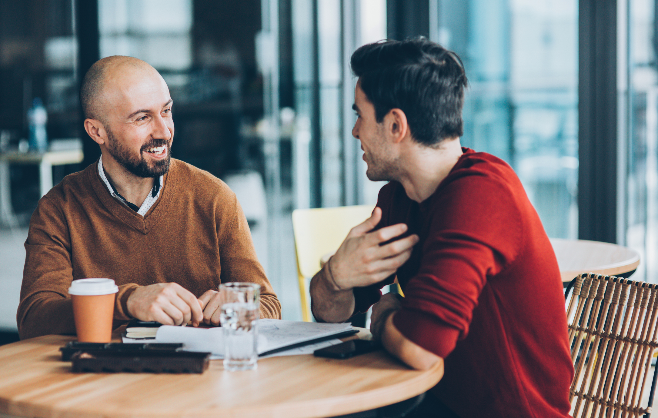 Two men are sitting at a table, engaged in a friendly conversation, with notebooks, a coffee cup, and a glass of water in front of them