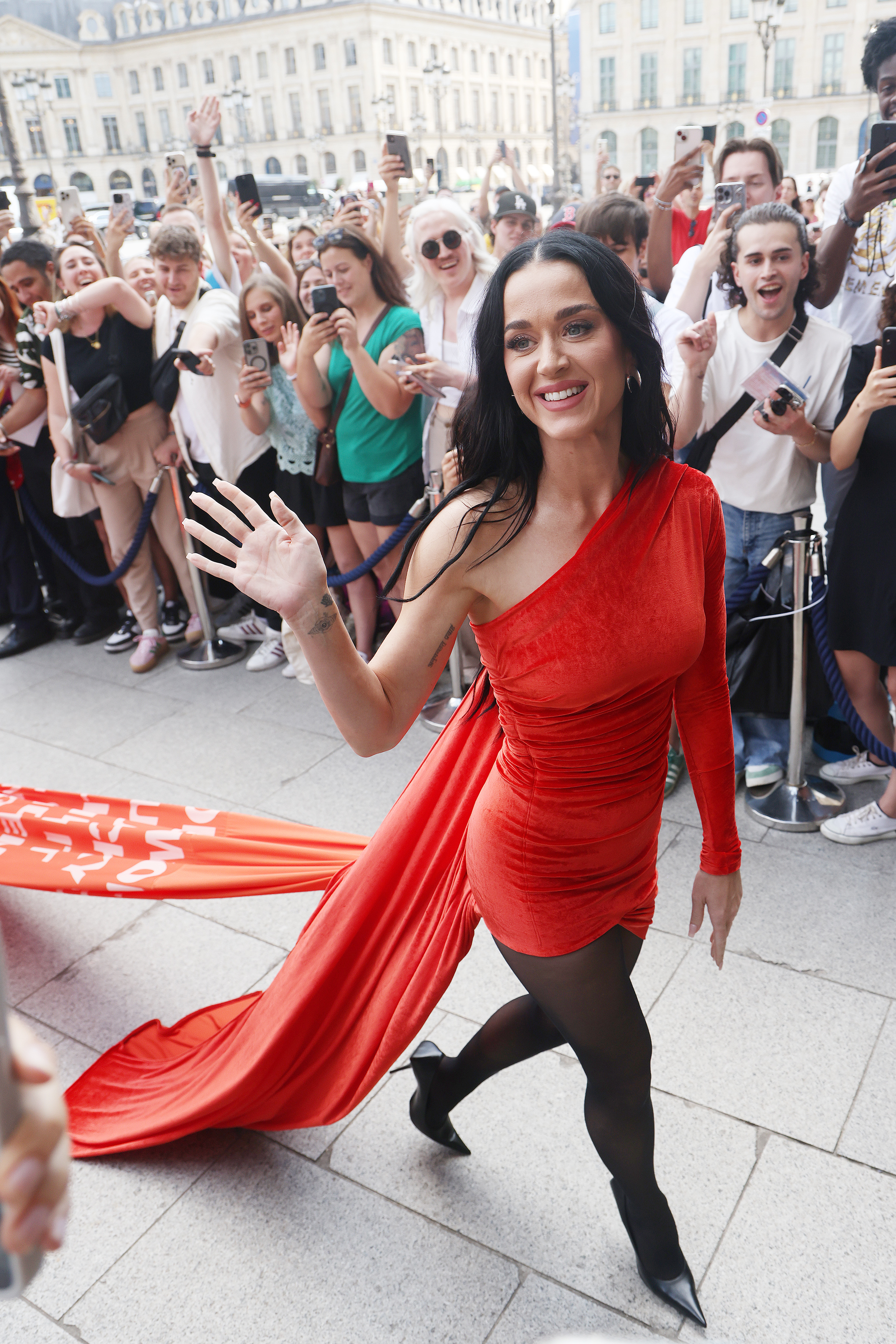 Katy Perry waves to a crowd while walking on a street in an elegant one-shoulder red dress with a long train, accompanied by black tights and heels