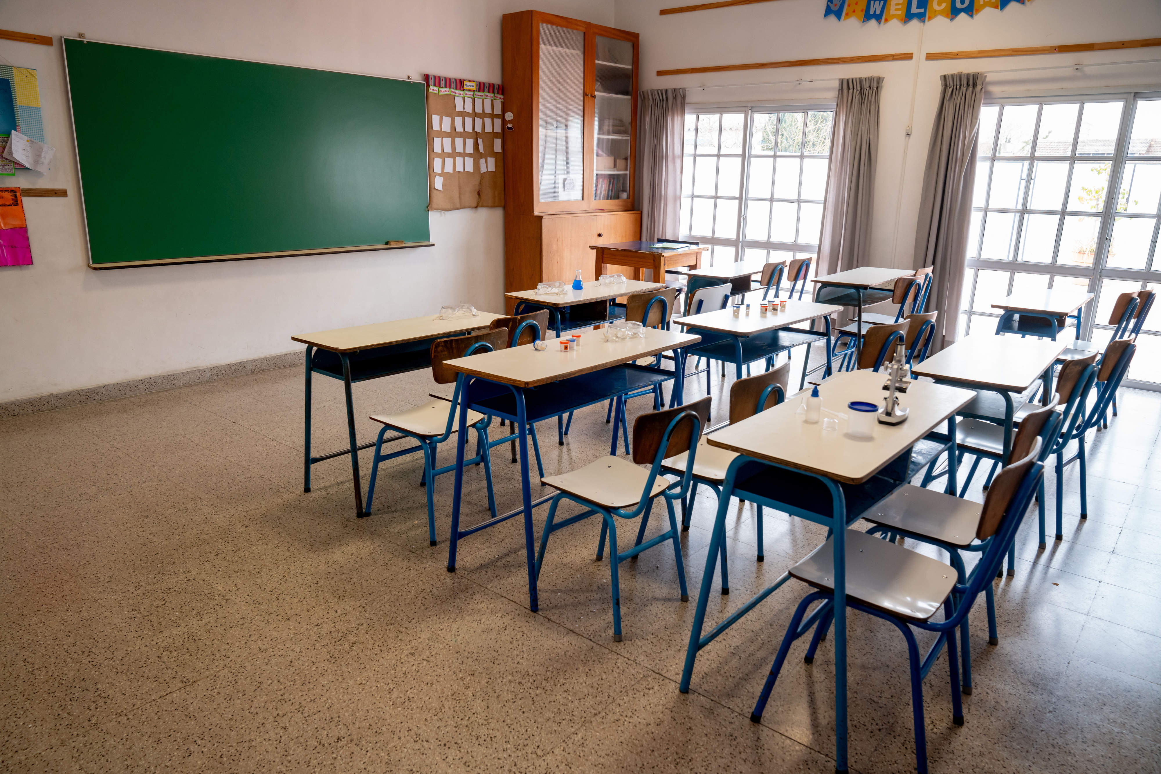 Classroom with empty desks and chairs, shelves with supplies, a green chalkboard, and large windows covered by curtains