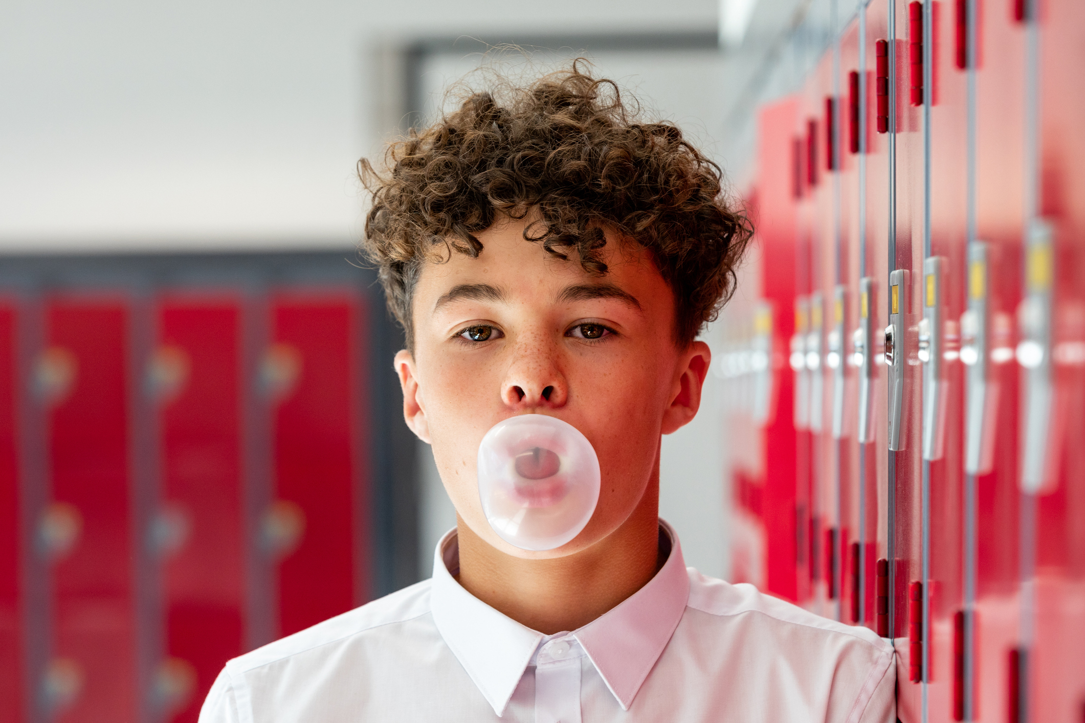 A young person with curly hair blows a bubble gum bubble in front of school lockers