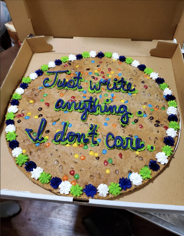 Large cookie cake with colorful icing that reads, &quot;Just write anything I don&#x27;t care,&quot; surrounded by small blue, green, white, and black icing decorations