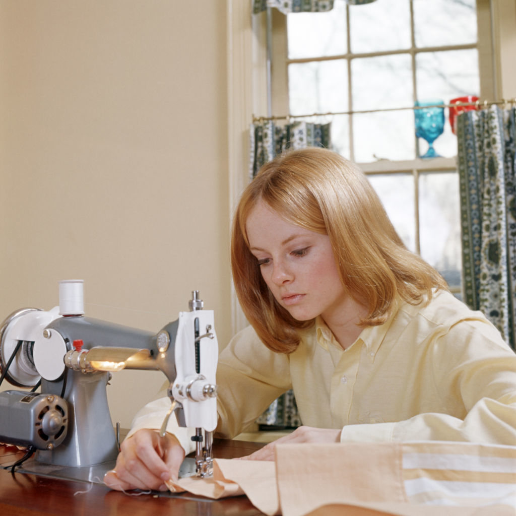 Woman focusing intently while sewing a garment on a sewing machine, possibly working from home. A window with curtains is visible in the background