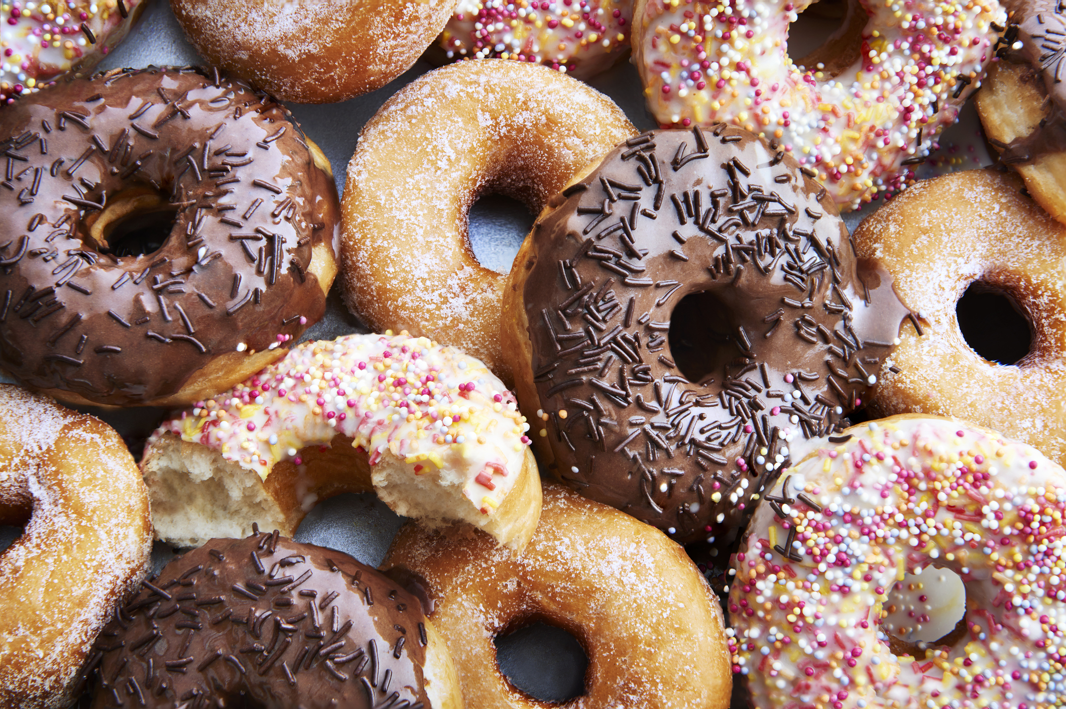 An assortment of donuts, some with chocolate glaze and sprinkles, others powdered or plain. One donut has a bite taken out of it