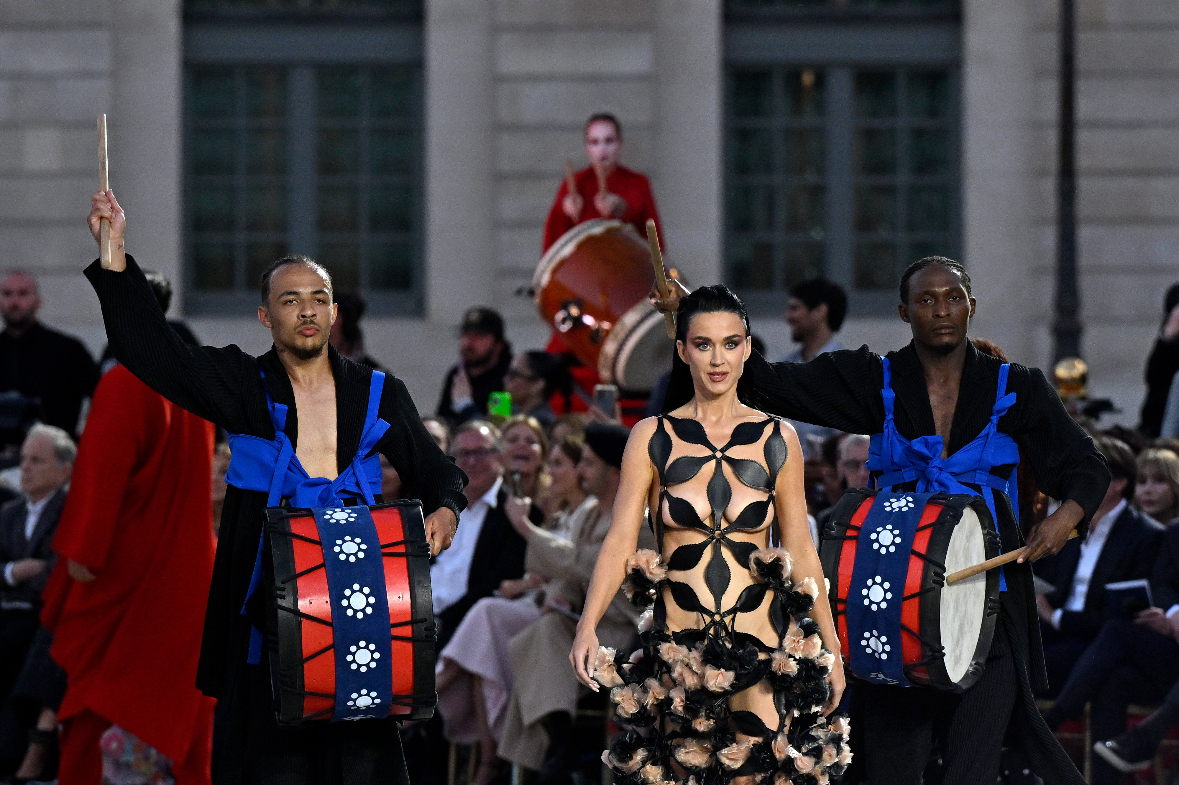 Katy Perry walks flanked by drummers. They perform in front of an audience seated in the background