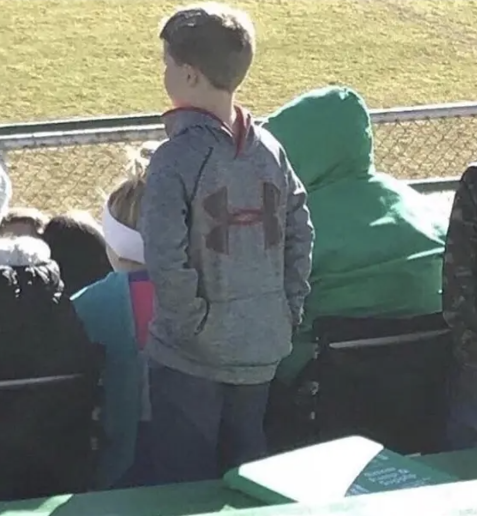 A young child in a hooded sweatshirt with a sports logo stands in a stadium, facing away, watching a game. Other spectators are seated nearby