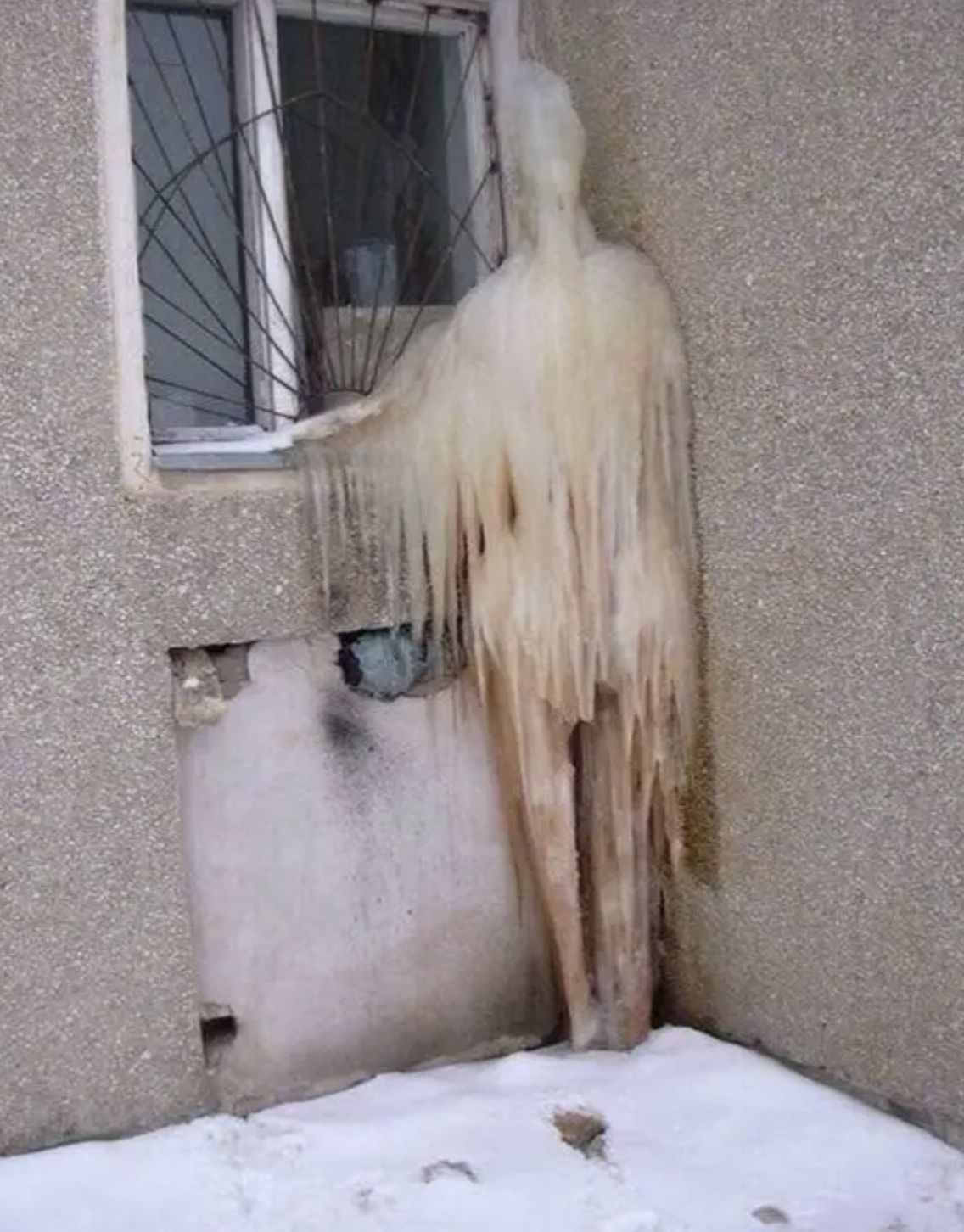 A frozen humanoid-like icicle formation drips from a window against a building. Snow covers the ground beneath