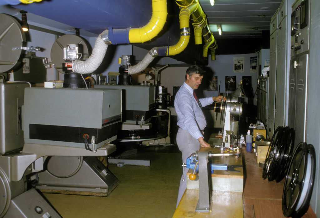 A man in a workroom operates large film equipment for projection or editing. Several machines and tools are visible on desks around him