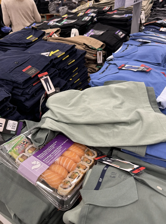 Casual men&#x27;s clothing like pants and shirts displayed on a store table, with a package of sushi placed on top of the clothes