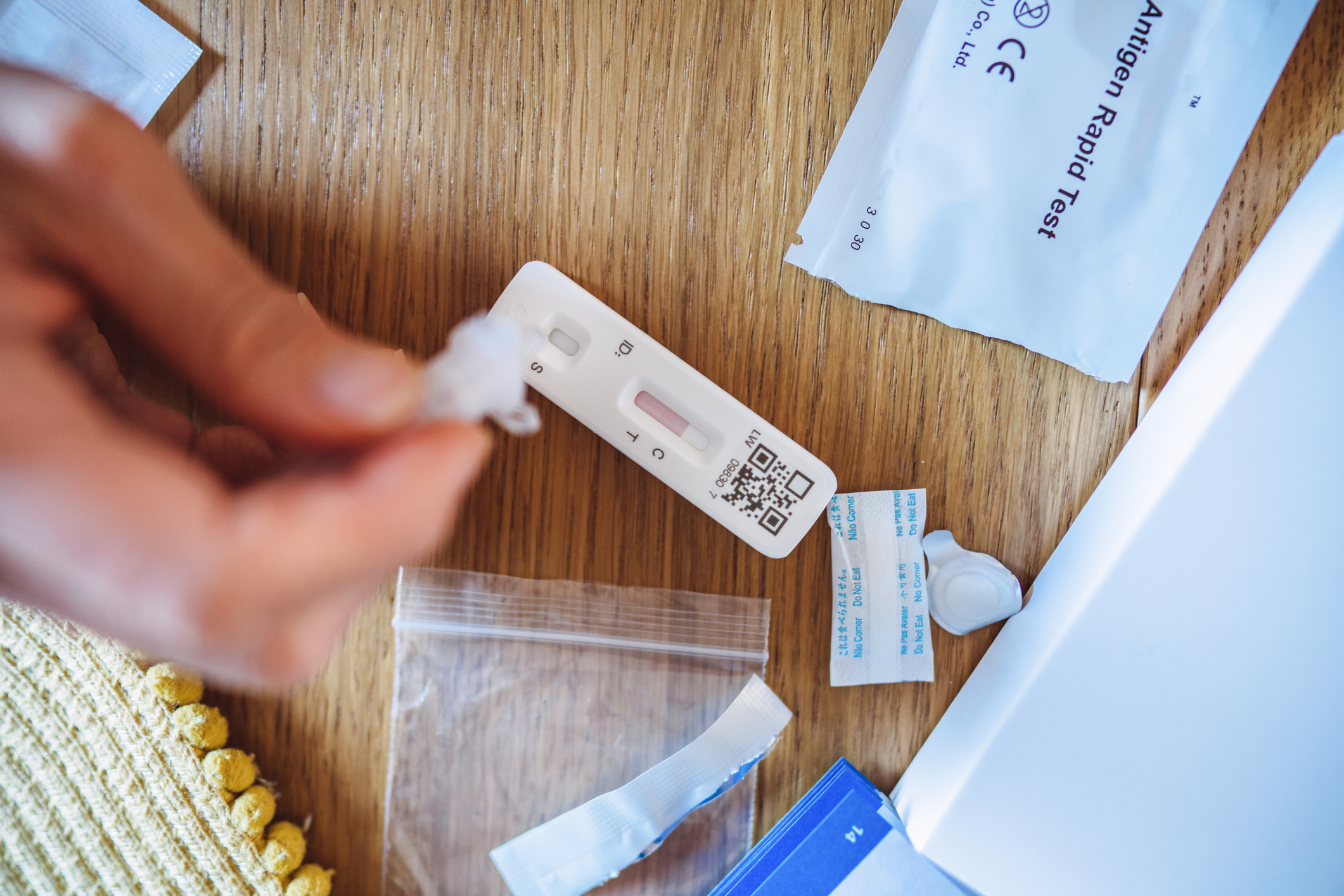 A hand holds a dropper over an at-home antigen rapid test on a wooden surface, surrounded by various components of the test kit