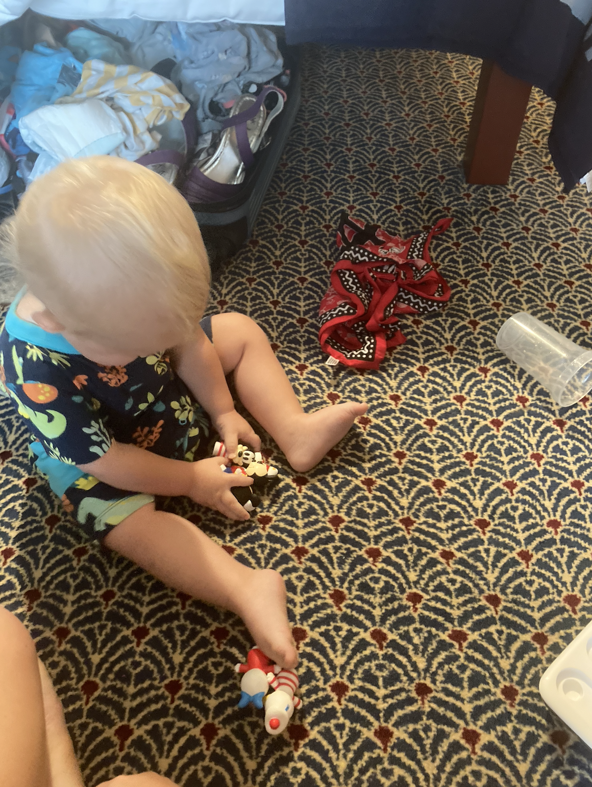A toddler sits on a patterned carpet playing with Disney toys, surrounded by scattered items near a bed