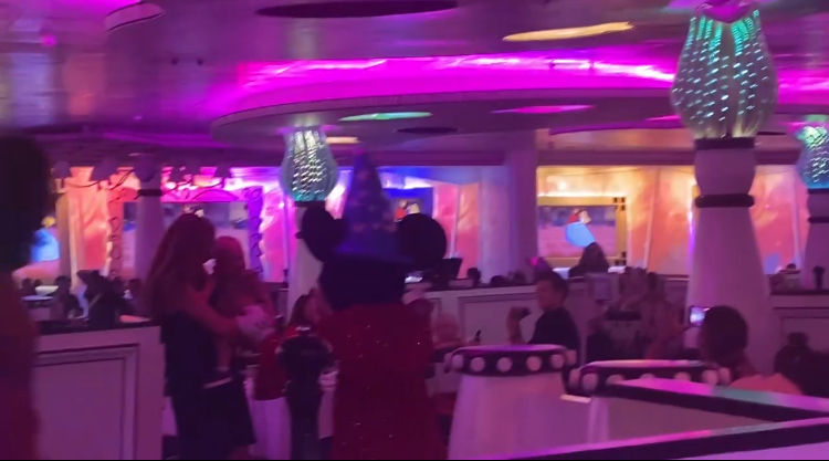 Mickey Mouse greets guests in a restaurant filled with animated decor and vibrant lighting. Diners are seated around tables, enjoying the lively atmosphere