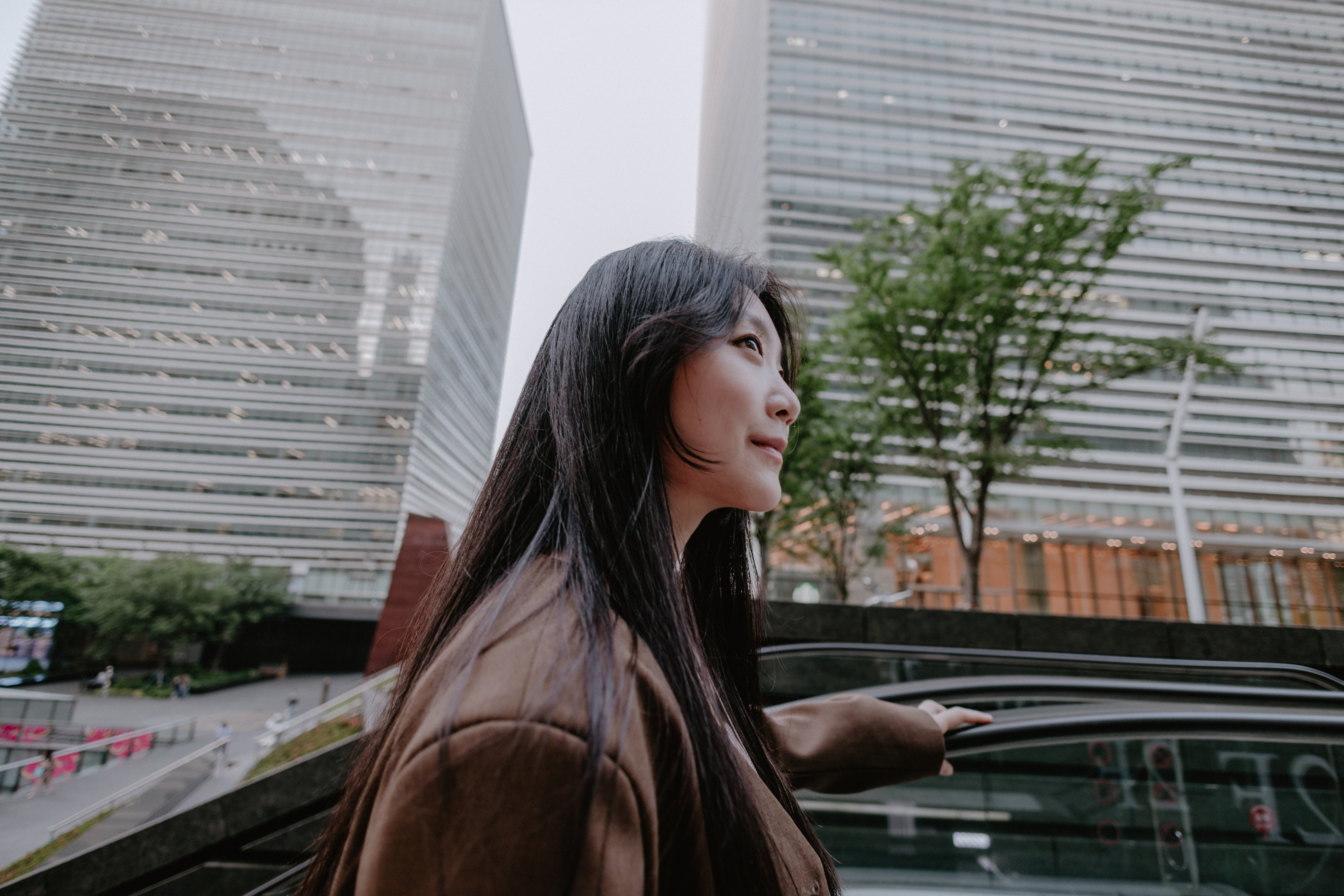 A person with long hair and a brown jacket stands outside, looking up at tall modern buildings and a tree in an urban setting