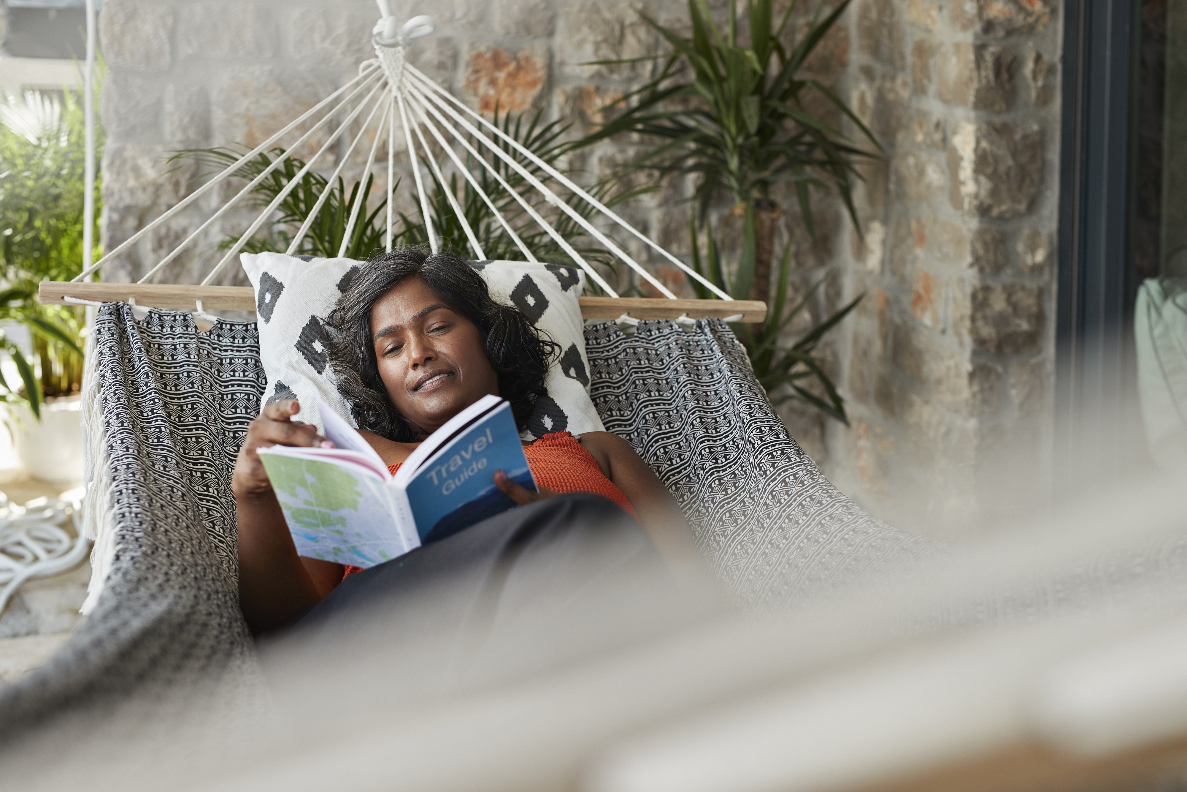 A woman is relaxing in a hammock, reading a book titled &quot;Travel.&quot; The setting is a cozy patio with some plants in the background