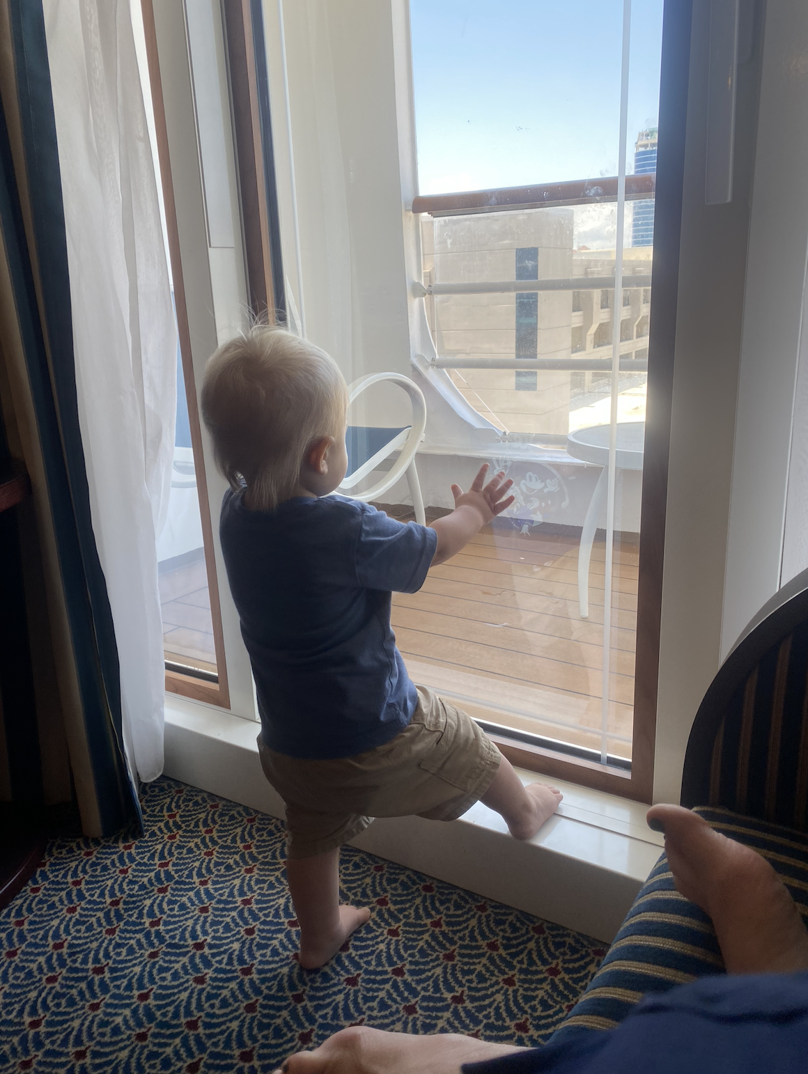 A toddler in a blue shirt and shorts, standing on a window sill, looking outside through a glass door at a balcony and distant buildings