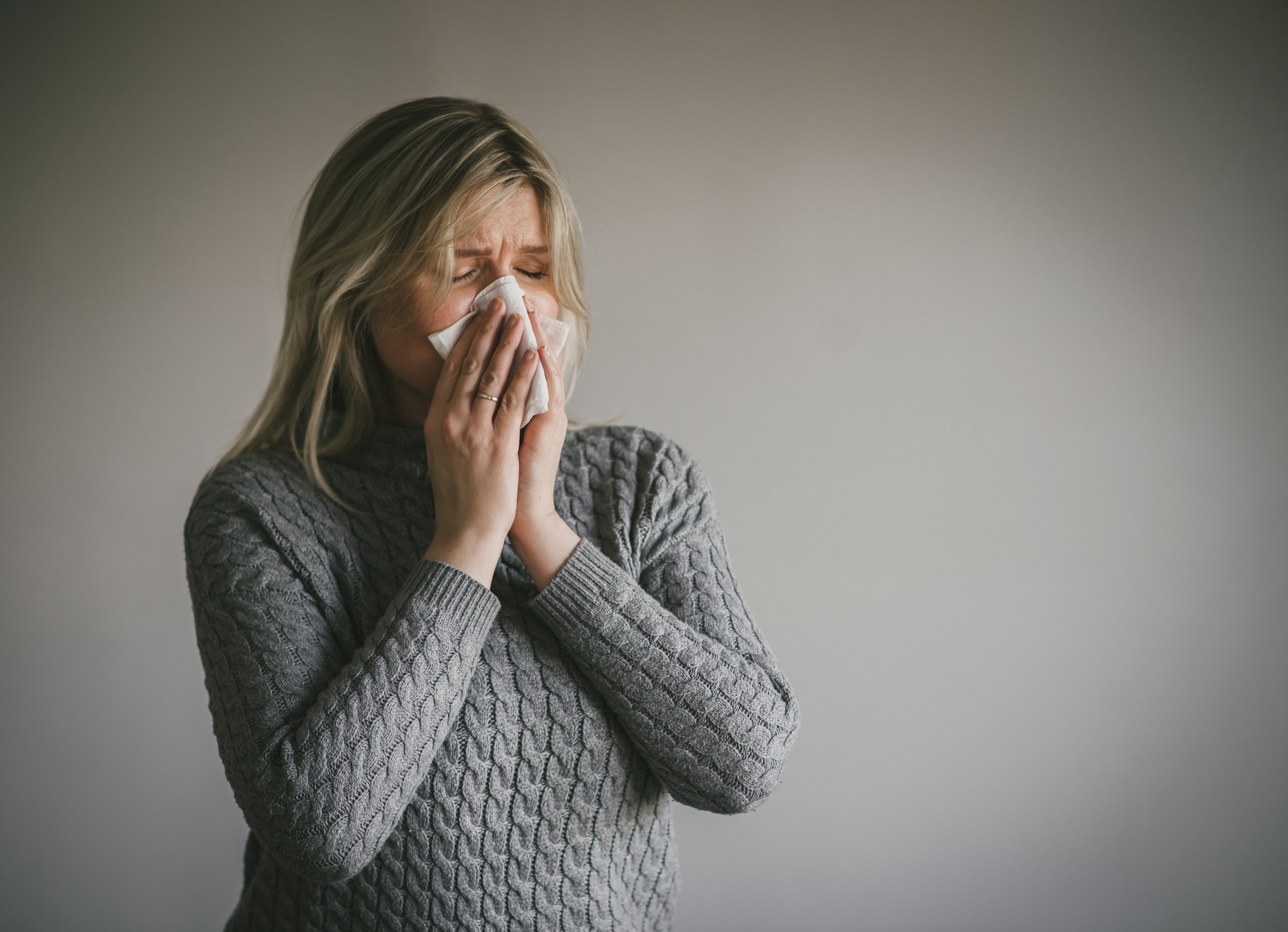 A person with long hair wearing a knit sweater is sneezing into a tissue, depicting symptoms of a cold or allergies