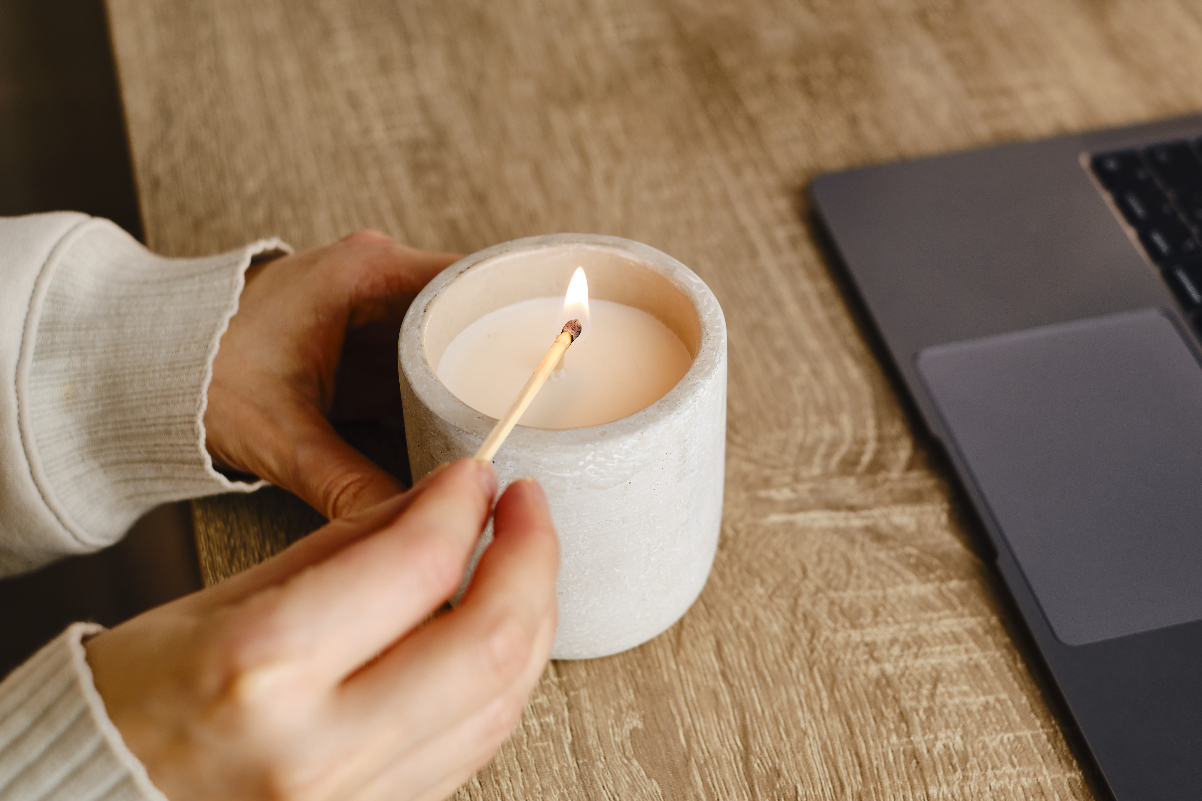 A person in a white sweater is lighting a candle on a wooden table next to a laptop