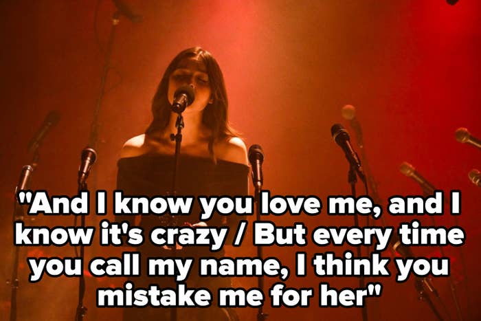 A female singer in an off-the-shoulder dress stands on stage, surrounded by microphones, performing under dramatic lighting. No identifying information available