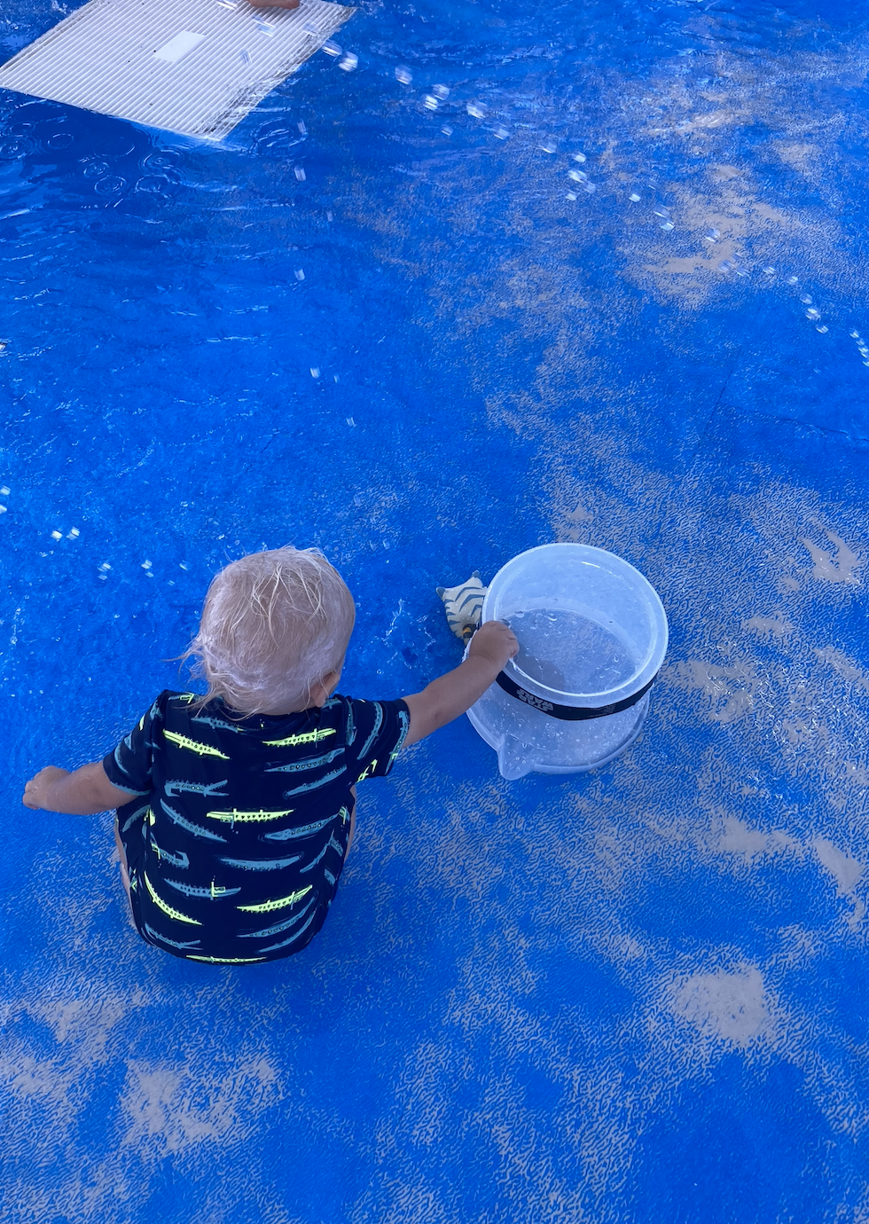 A toddler sits on the ground, wearing a printed shirt, reaching towards a clear bucket of water on a blue textured surface