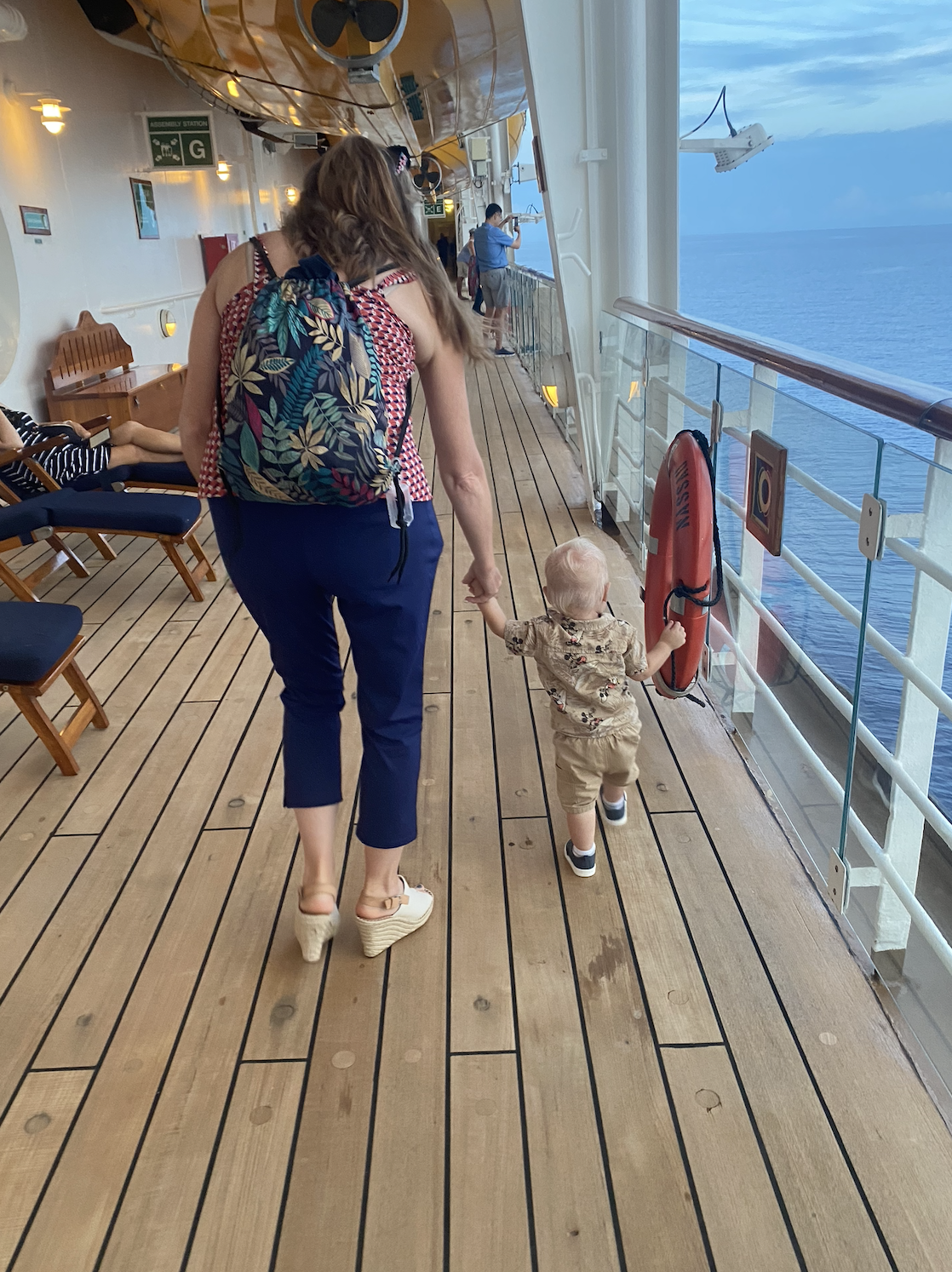 Woman holding hands with a child while walking on the deck of a cruise ship. The woman is wearing cropped pants and a patterned sleeveless top. The child is dressed in a printed outfit