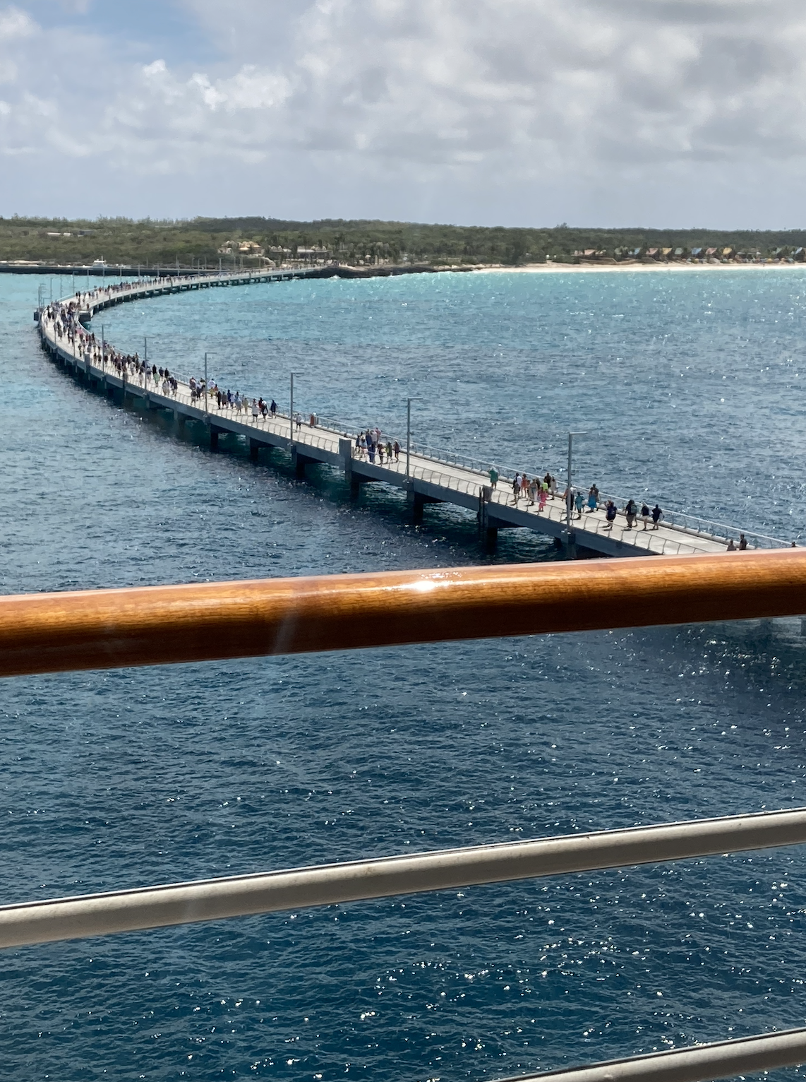 View of a curvy pier extending into the ocean with people walking along it. Farmland and buildings are visible in the distant background