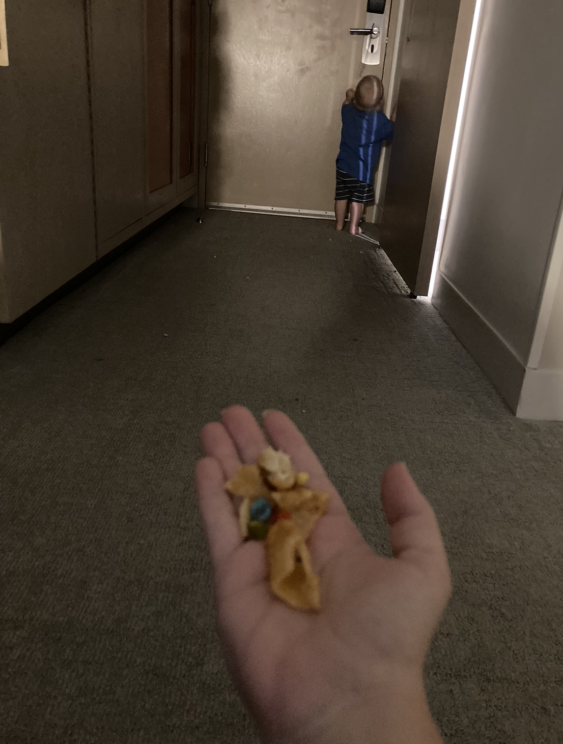 A child wearing a blue jacket is at the end of a hallway, trying to open a door. An adult hand in the foreground holds snack pieces