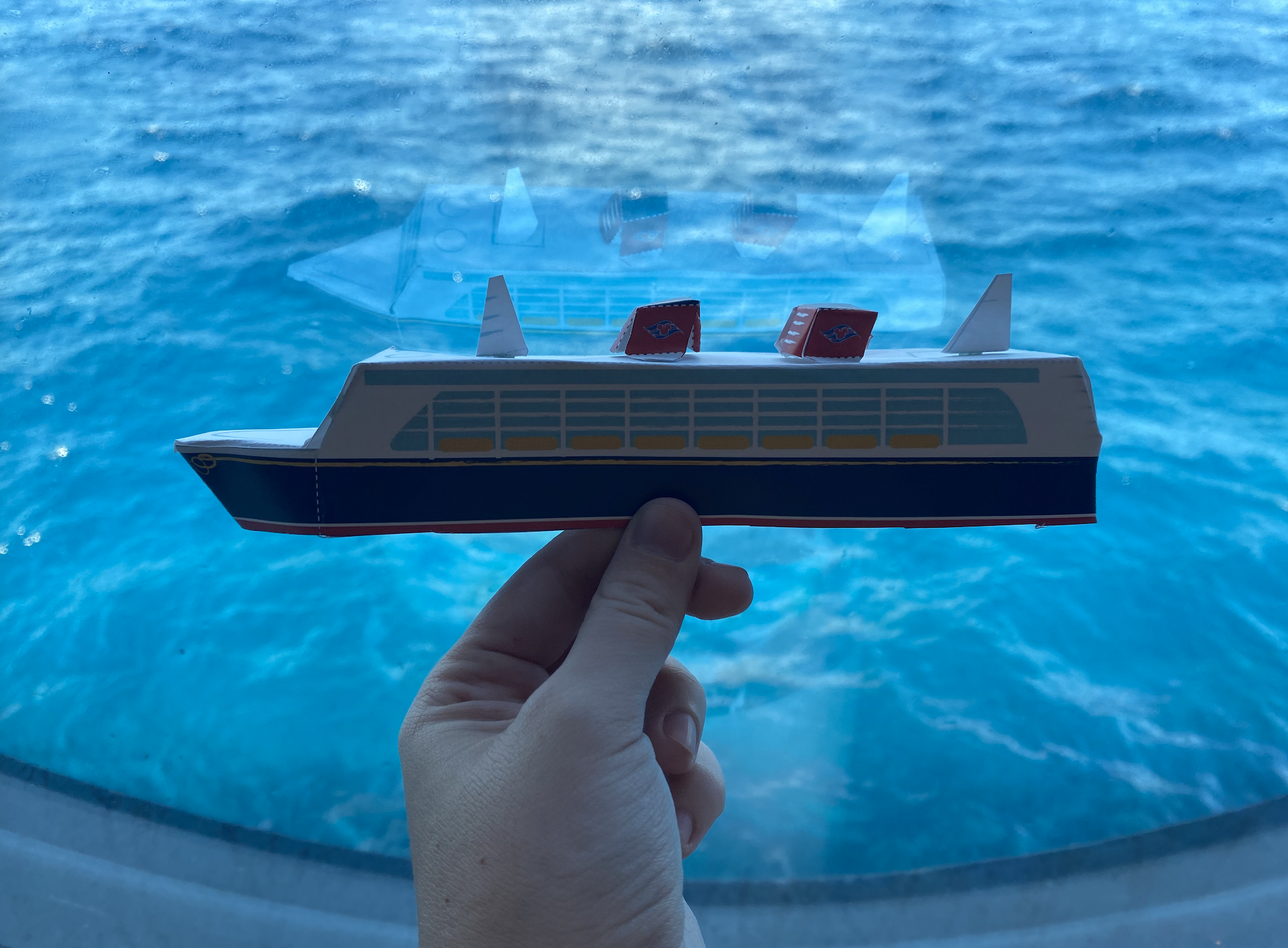 A hand holding a small paper model of a cruise ship in front of a window showing the ocean water outside