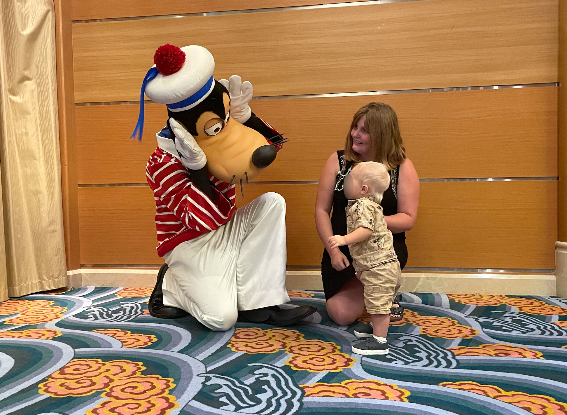 Goofy in a sailor costume playfully interacts with a woman and a toddler on a carpeted floor