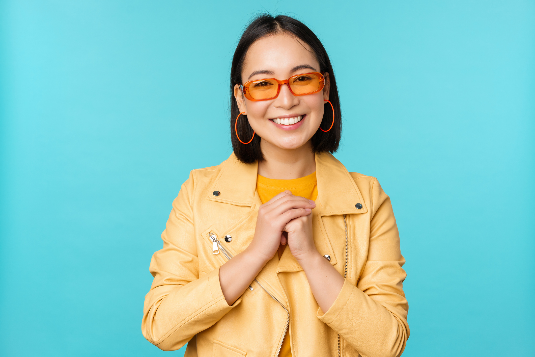 A smiling woman wearing a yellow jacket, orange-tinted glasses, and hoop earrings stands against a plain background, with hands clasped in front
