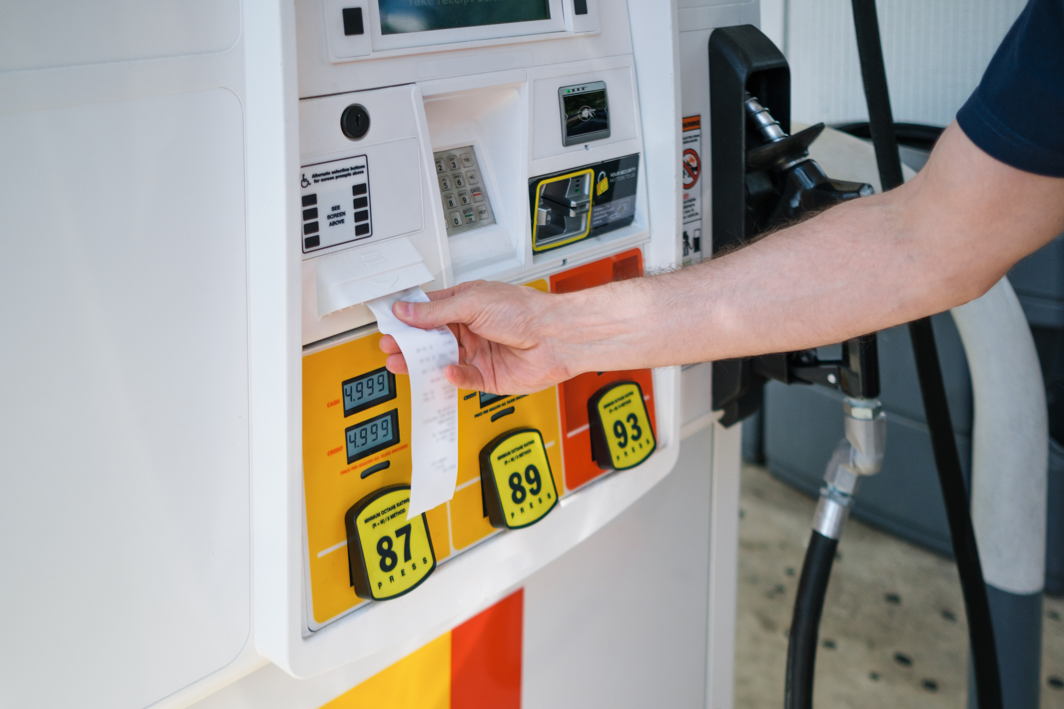 A person retrieves a printed receipt from a gas pump after filling up their vehicle. The gas prices for octane levels 87, 89, and 93 are displayed