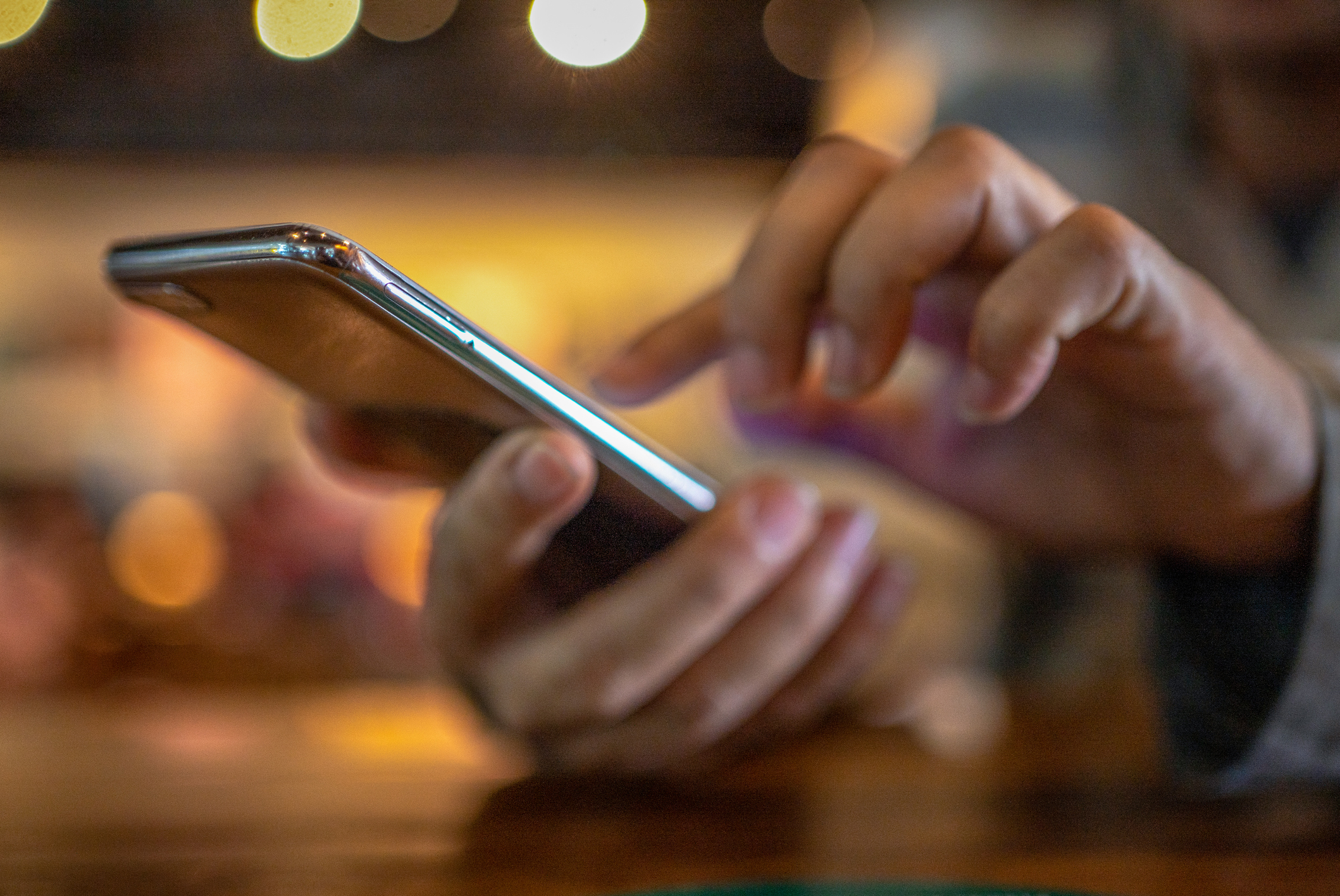 Close-up of a person&#x27;s hands holding and using a smartphone. The background is blurred, with small lights visible