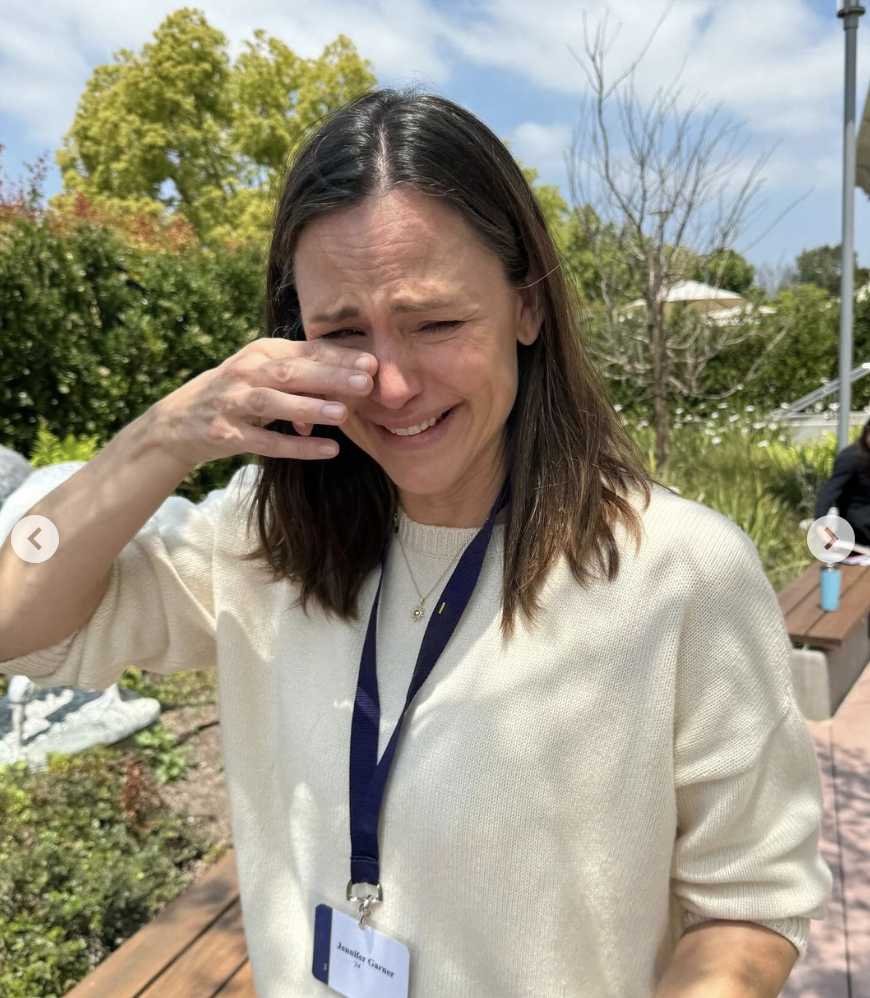 Jennifer Garner appears to be emotional while wiping her eye, wearing a white sweater and a lanyard with a name tag. Outdoor setting with greenery in the background